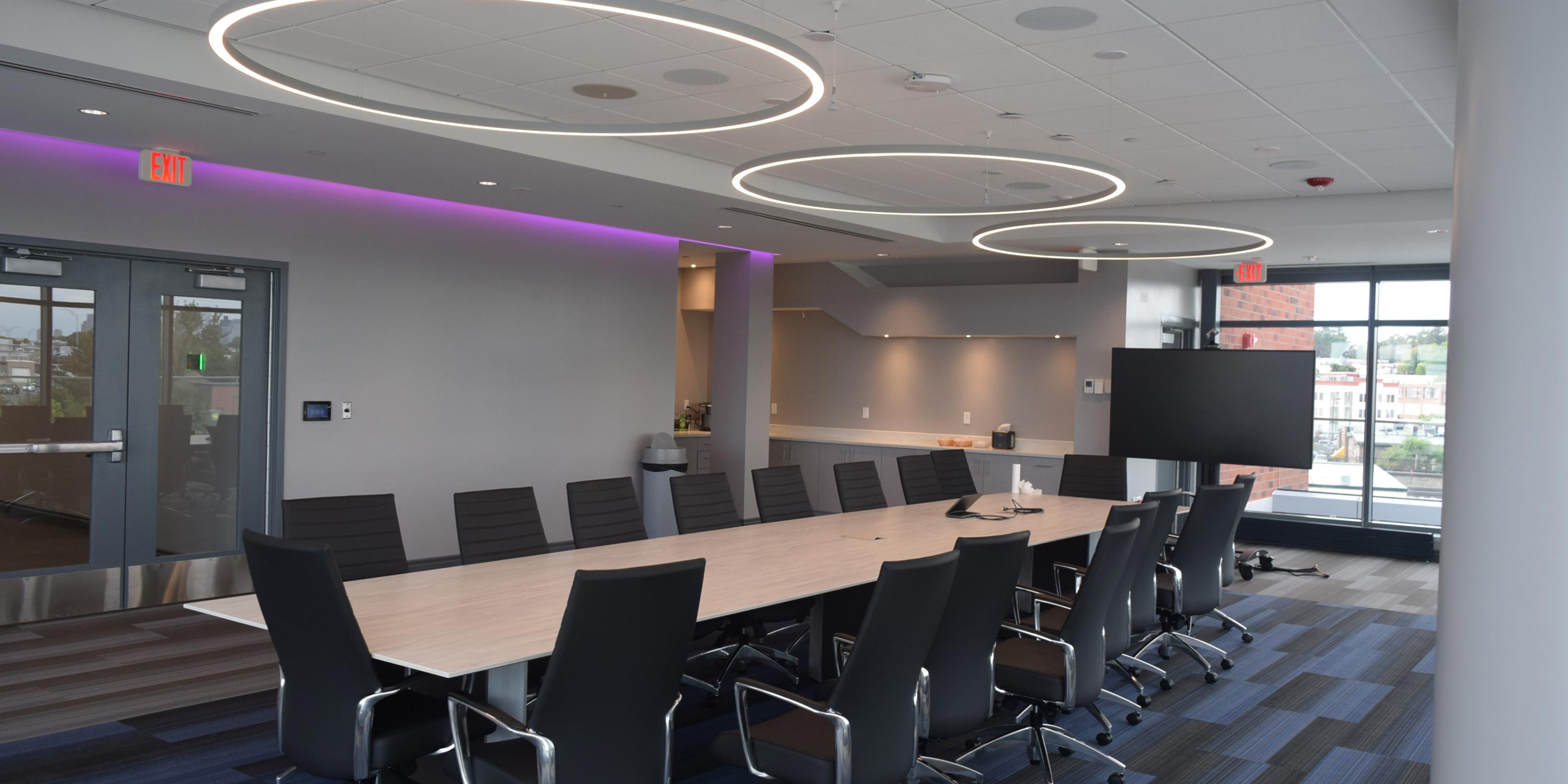 A sleek conference room with a long table, black chairs, and circular ceiling lights overhead.