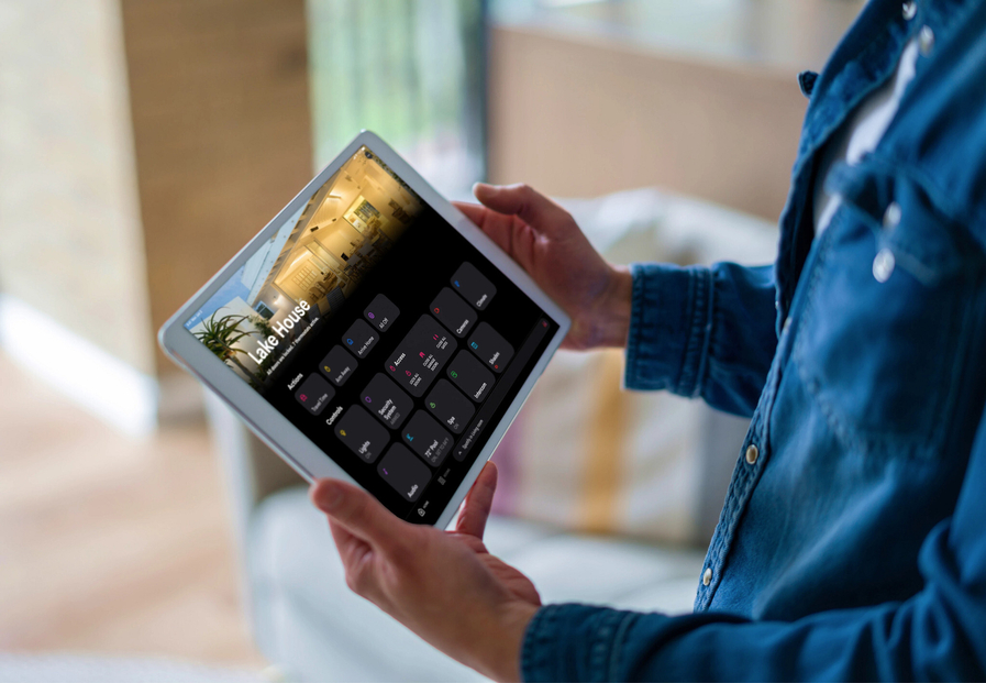 Person holding a tablet that displays the Crestron home control interface, with a screen labeled “Lake House.”