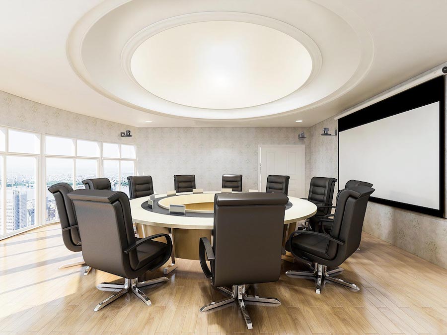 A modern conference room with high-back chairs, a large round table, and a ceiling-mounted projector screen, surrounded by floor-to-ceiling windows with city views.