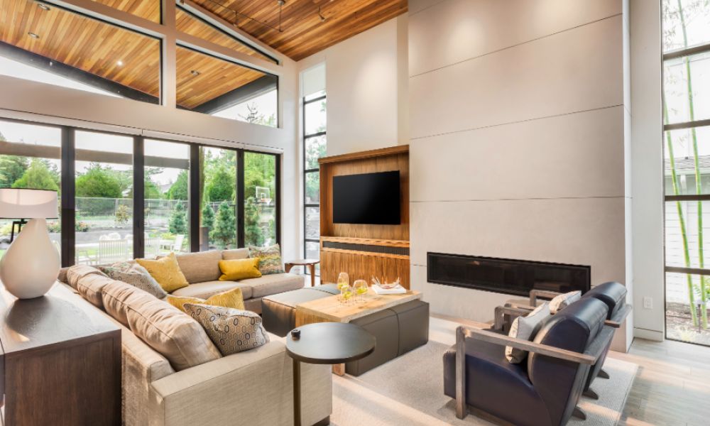 A contemporary living room featuring large windows, a wood-paneled ceiling, a wall-mounted TV, modern furnishings, and a linear gas fireplace.
