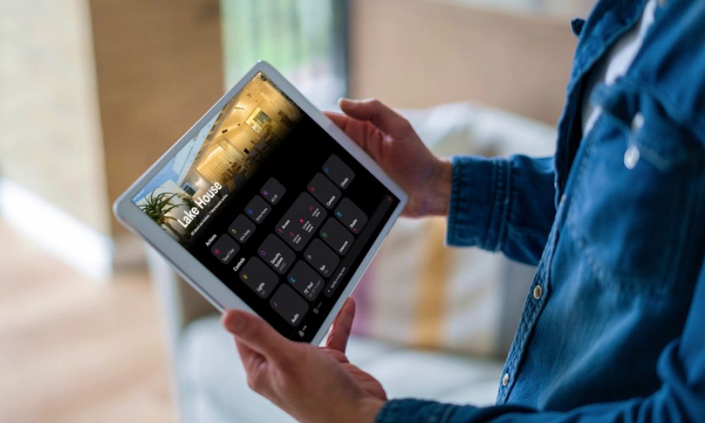 Person holding a tablet that displays the Crestron home control interface, with a screen labeled “Lake House.”